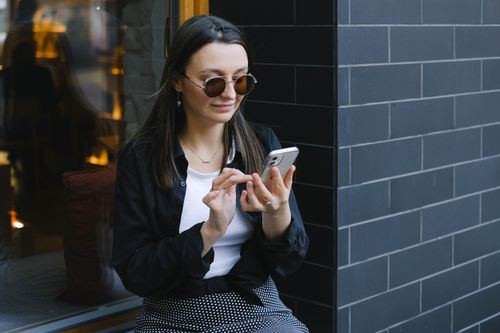 Free Trendy woman with sunglasses using a smartphone outdoors. Urban lifestyle captured in a candid moment. Stock Photo