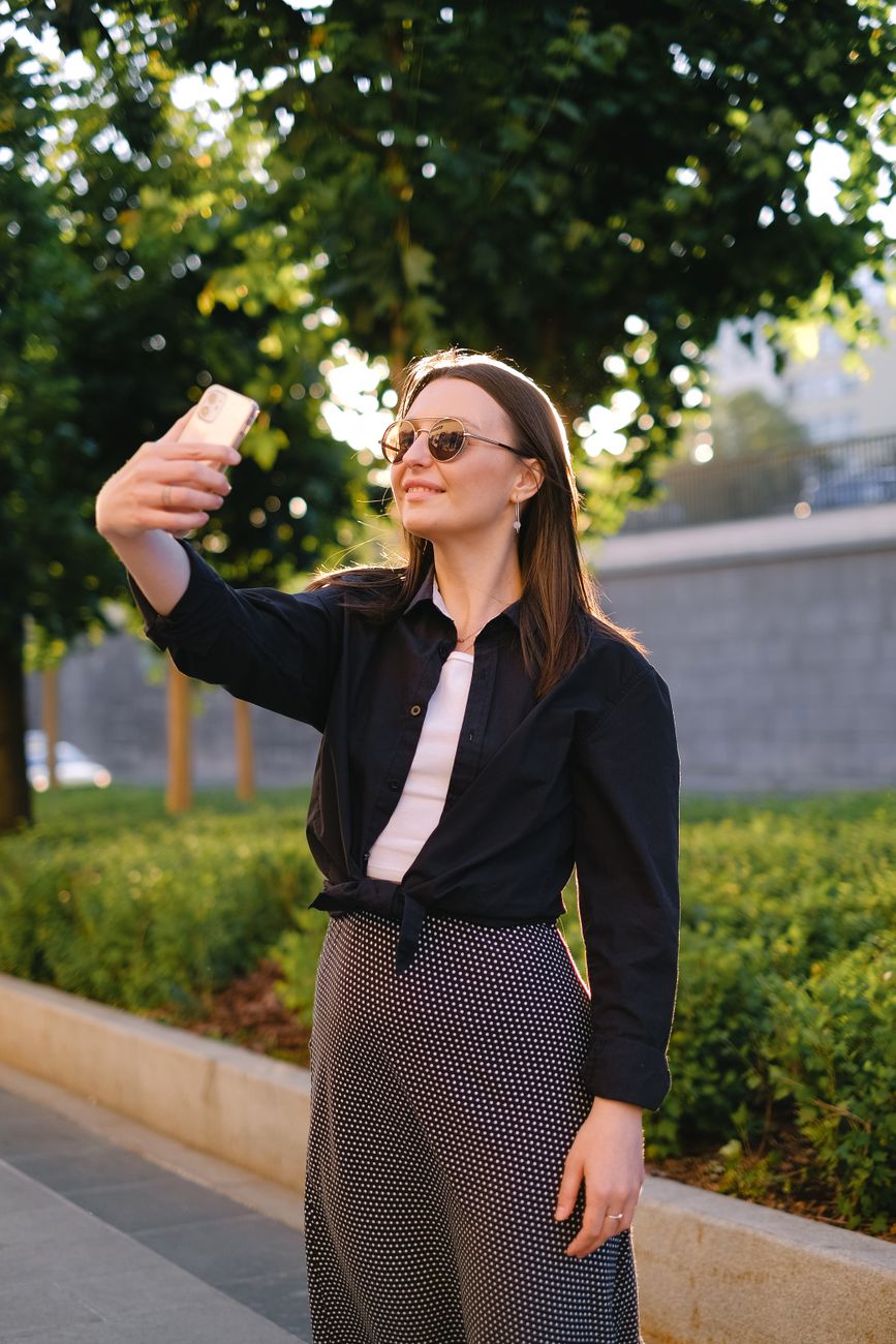 Smiling woman taking a selfie with smartphone outdoors in sunlight wearing sunglasses and fashionabl