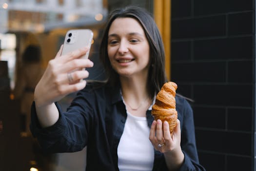 Smiling woman holding croissant while taking a selfie with a smartphone outdoors.