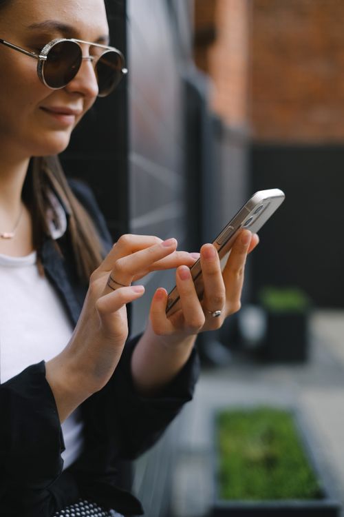 Free Woman holding smartphone, using it outdoors on a sunny day, wearing sunglasses. Stock Photo
