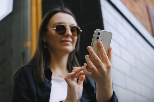 Free A young woman using a smartphone outdoors, wearing sunglasses and smiling confidently. Stock Photo