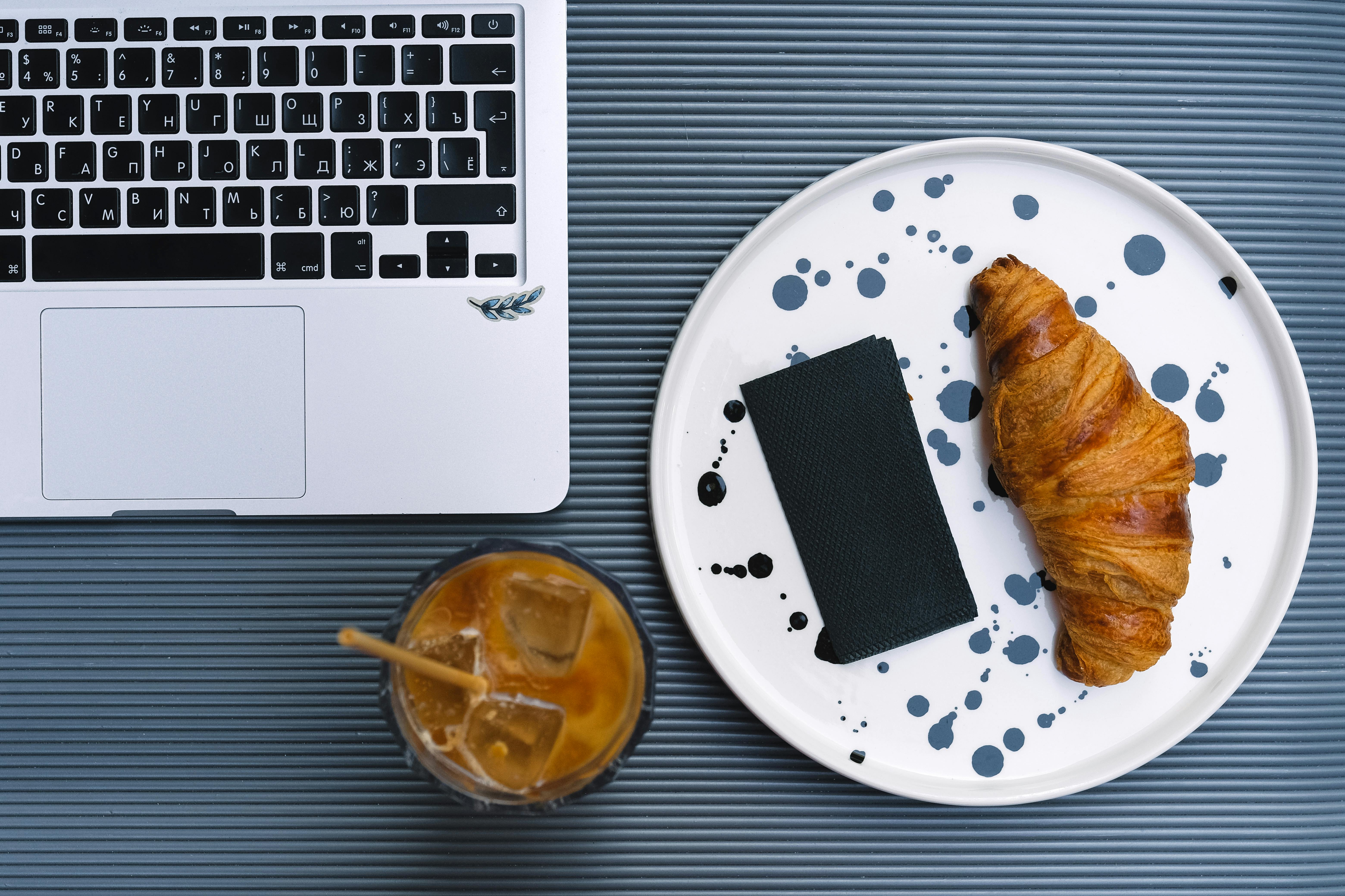 A stylish flat lay of a croissant, iced latte, and laptop, perfect for work-from-home inspiration.