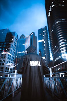 Person in hooded coat overlooks illuminated skyscrapers in city at night. Modern urban vibe.