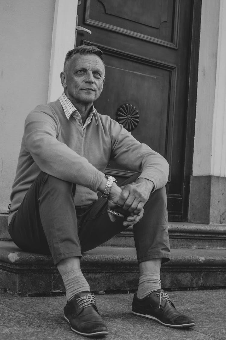 Man In Long Sleeve Shirt And Pants Sitting At A Doorstep