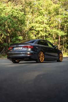 A stylish black sedan with golden rims parked on a forest-lined road in daylight.