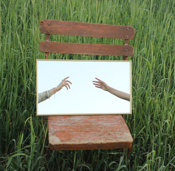 Mirror on a wooden chair reflects hands reaching in a grassy field, creating a surreal outdoor scene.