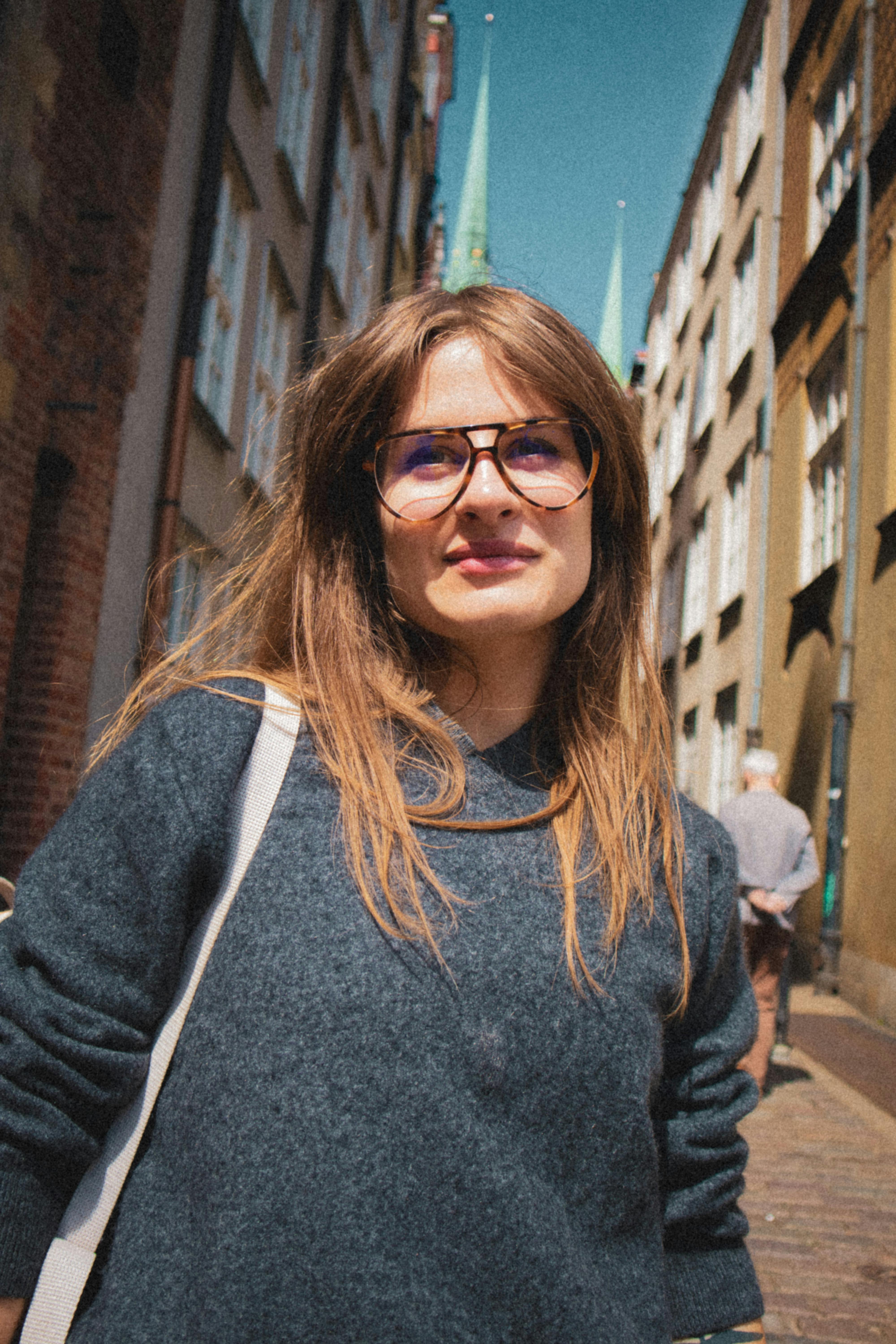 Confident woman with glasses smiling on a sunny street, surrounded by urban architecture.