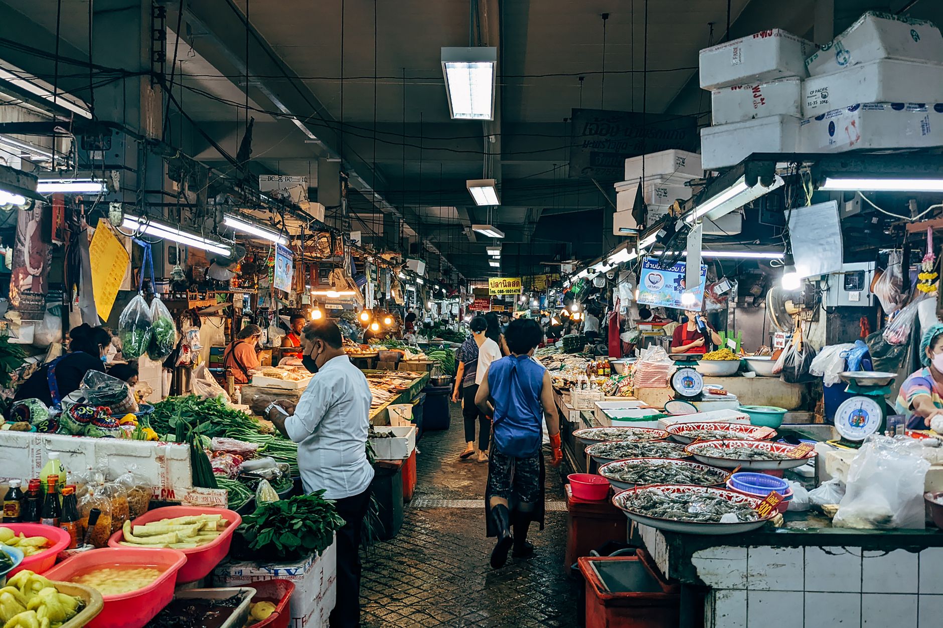 A vibrant scene at an indoor wet market with various fresh produce and shoppers browsing stalls.