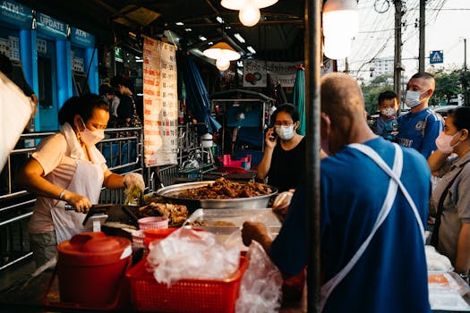 Market vendor selling food at a street market in Bangkok, with people wearing face masks.