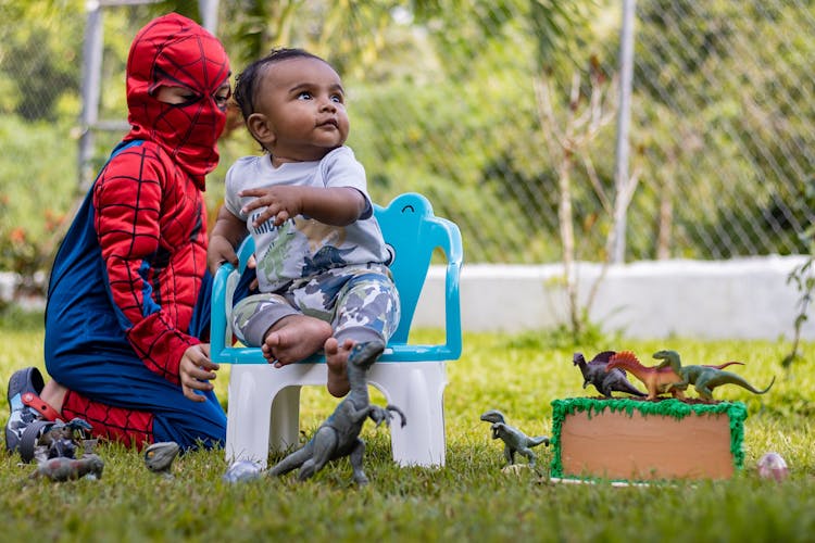 A Toddler Sitting On A Chair Beside A Kid In Spiderman Costume