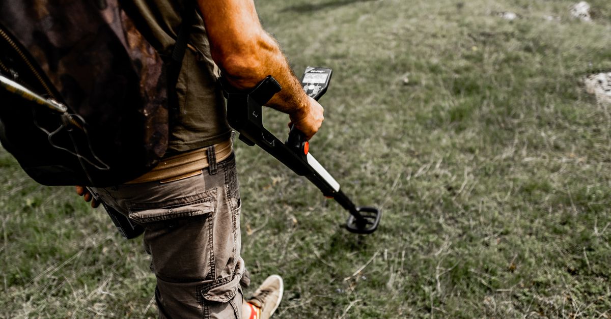 Person using a metal detector on a grassy field wearing casual attire.