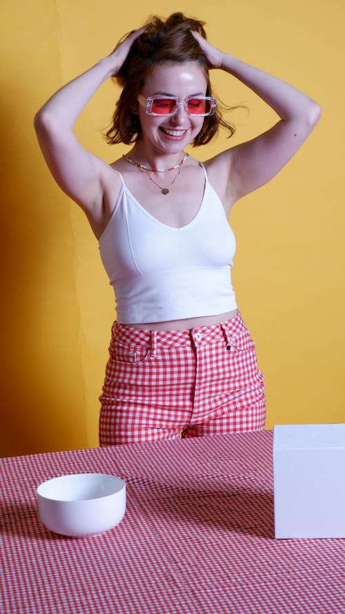 A Woman in White Tank Top and Red Checkered Shorts Smiling with Her Hands on Her Head