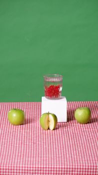 A colorful still life composition featuring green apples and a glass of red berries on a checkered tablecloth.