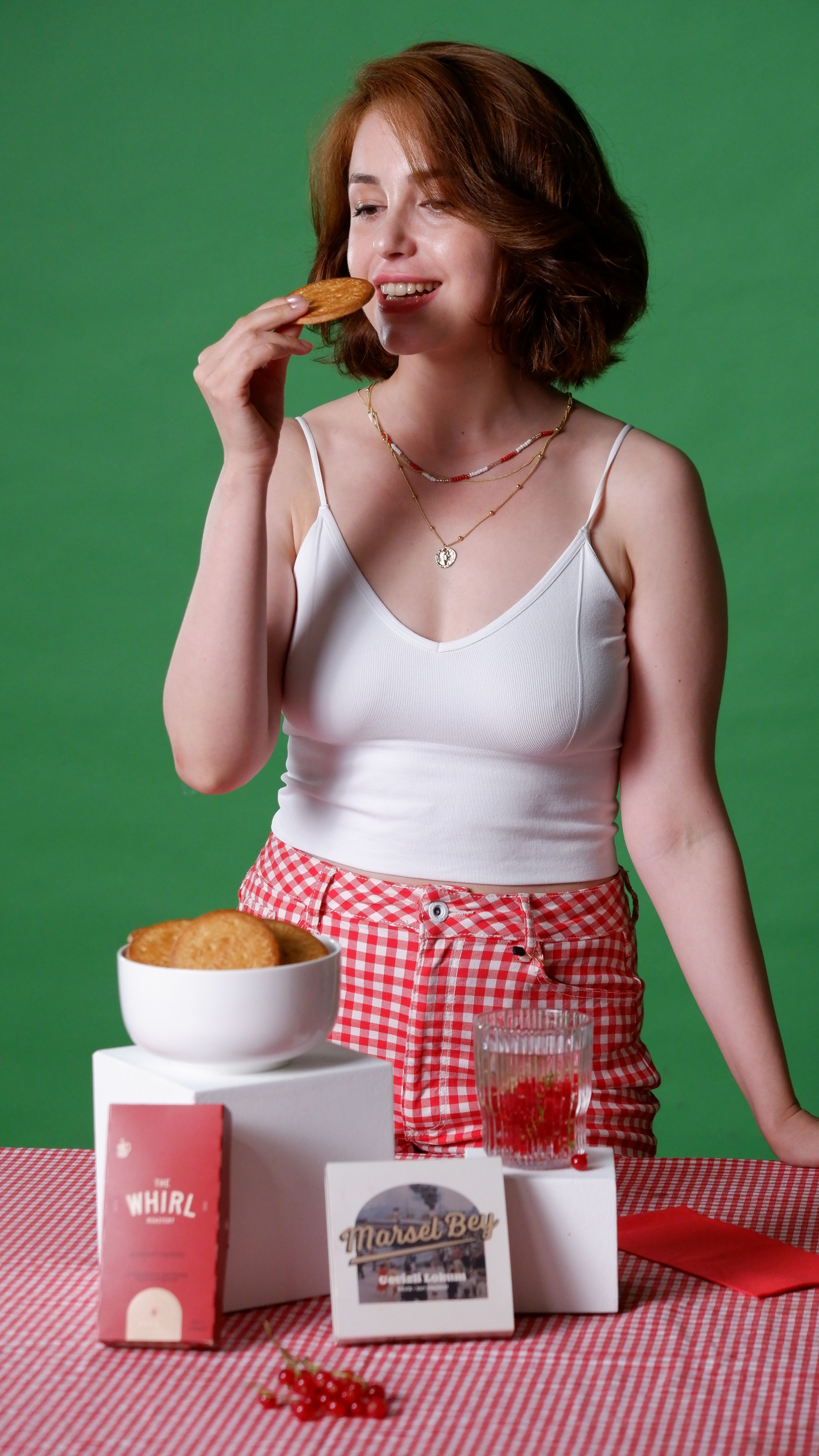Woman in white tank top enjoys cookies, standing by a table with red checkered cloth and a green background.