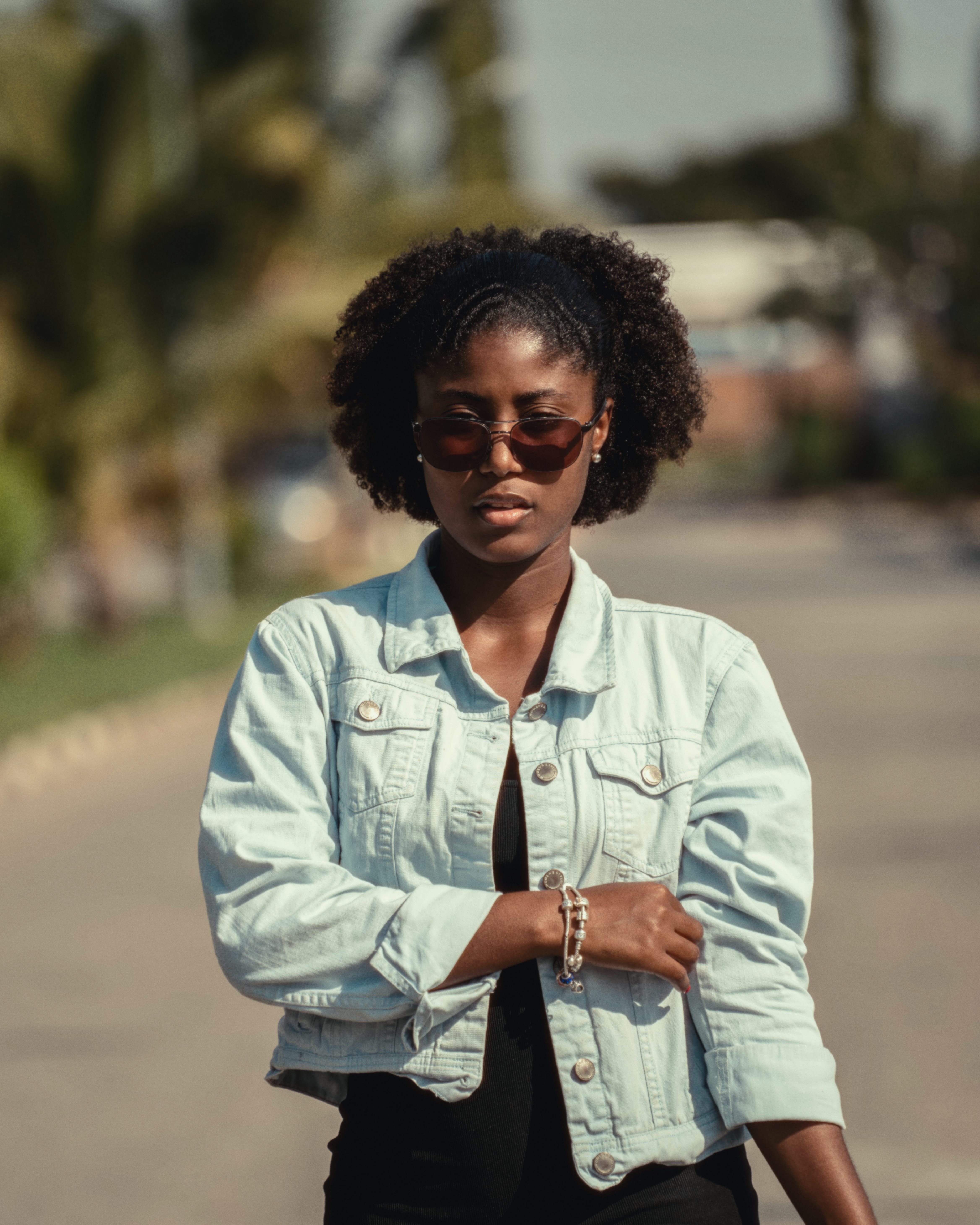 Portrait of a fashionable young woman wearing sunglasses and a denim jacket outdoors on a sunny day.