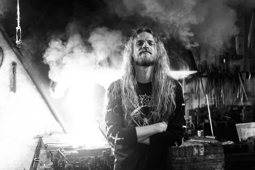 Dramatic black and white portrait of a long-haired male blacksmith in a workshop filled with smoke.