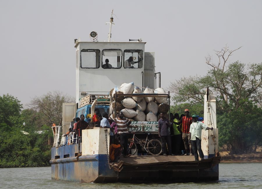 A ferry with passengers and goods travels across a river in Gambia, surrounded by lush greenery.