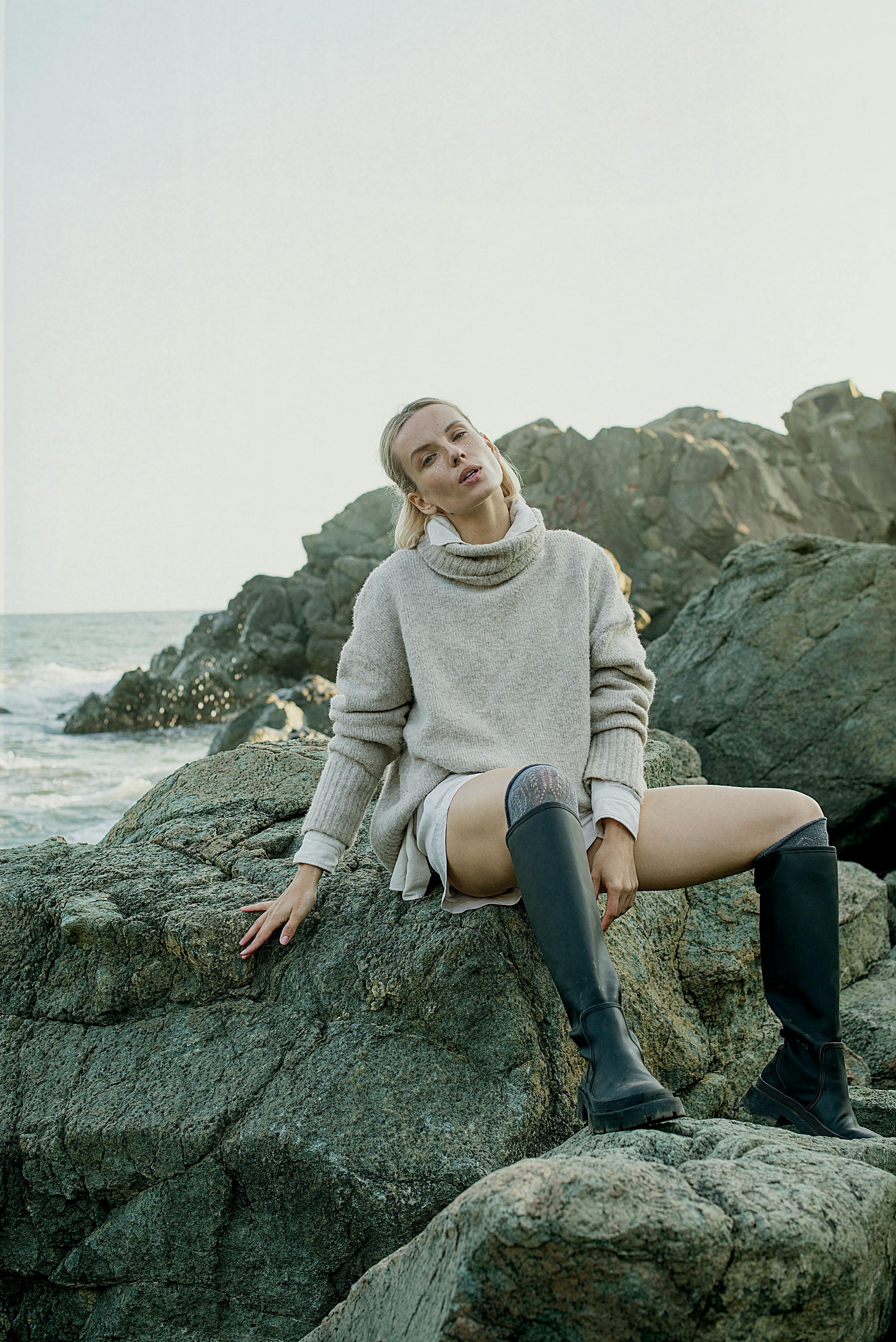 Blonde woman in a cozy sweater seated on coastal rocks, embracing nature.