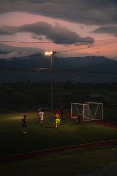 Players enjoy a soccer game at dusk under bright stadium lights and a colorful sky.