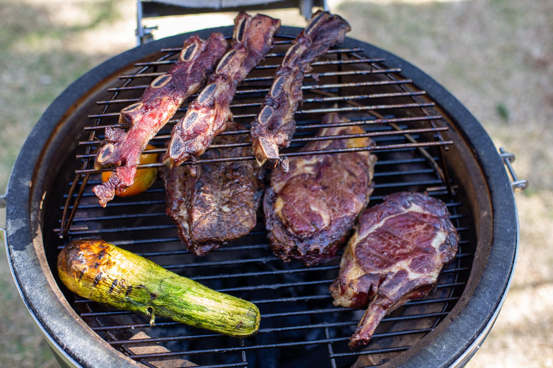 Close-up of assorted meats and zucchini grilling outdoors in daylight.