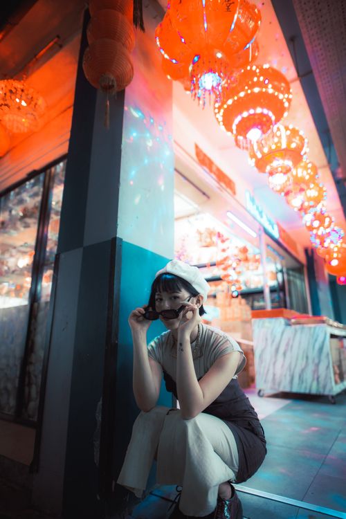 Free Young woman in a beige dress and sunglasses posing under illuminated lanterns on a vibrant street. Stock Photo