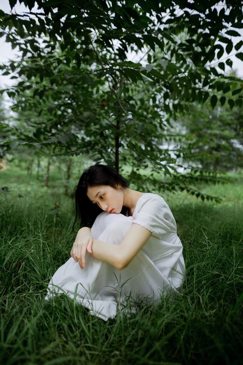 Free Relaxed Asian woman in white dress sitting on grass beneath a tree in a lush green field. Stock Photo
