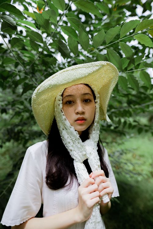 Free A young Asian woman wearing a straw hat stands amidst lush green foliage, creating a serene outdoor portrait. Stock Photo