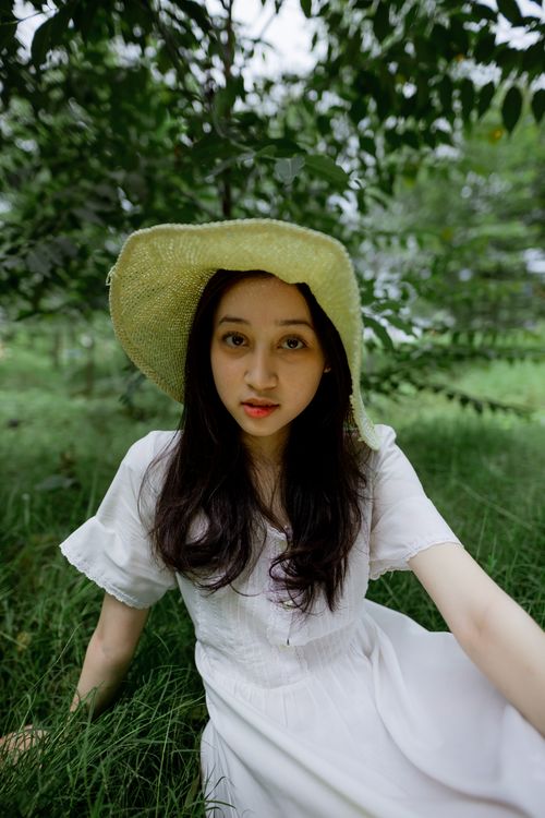 Free Asian woman in a white dress with a straw hat relaxing in a green park, capturing a serene summer moment. Stock Photo