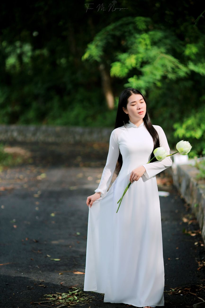 A serene portrait of a woman in a white dress holding flowers in a lush park setting.