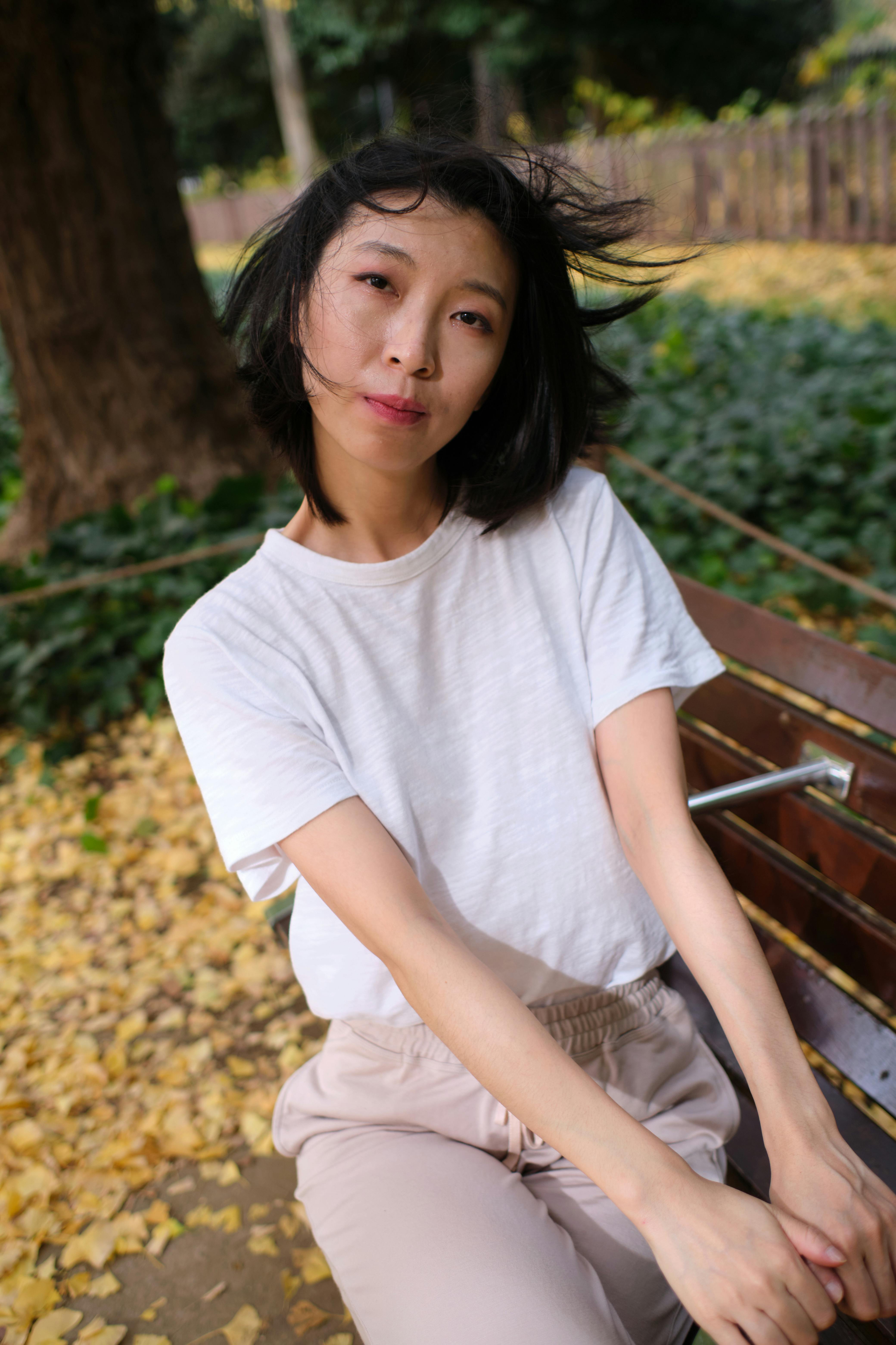 Asian woman sitting on a park bench amidst fallen leaves, capturing a serene autumn moment.