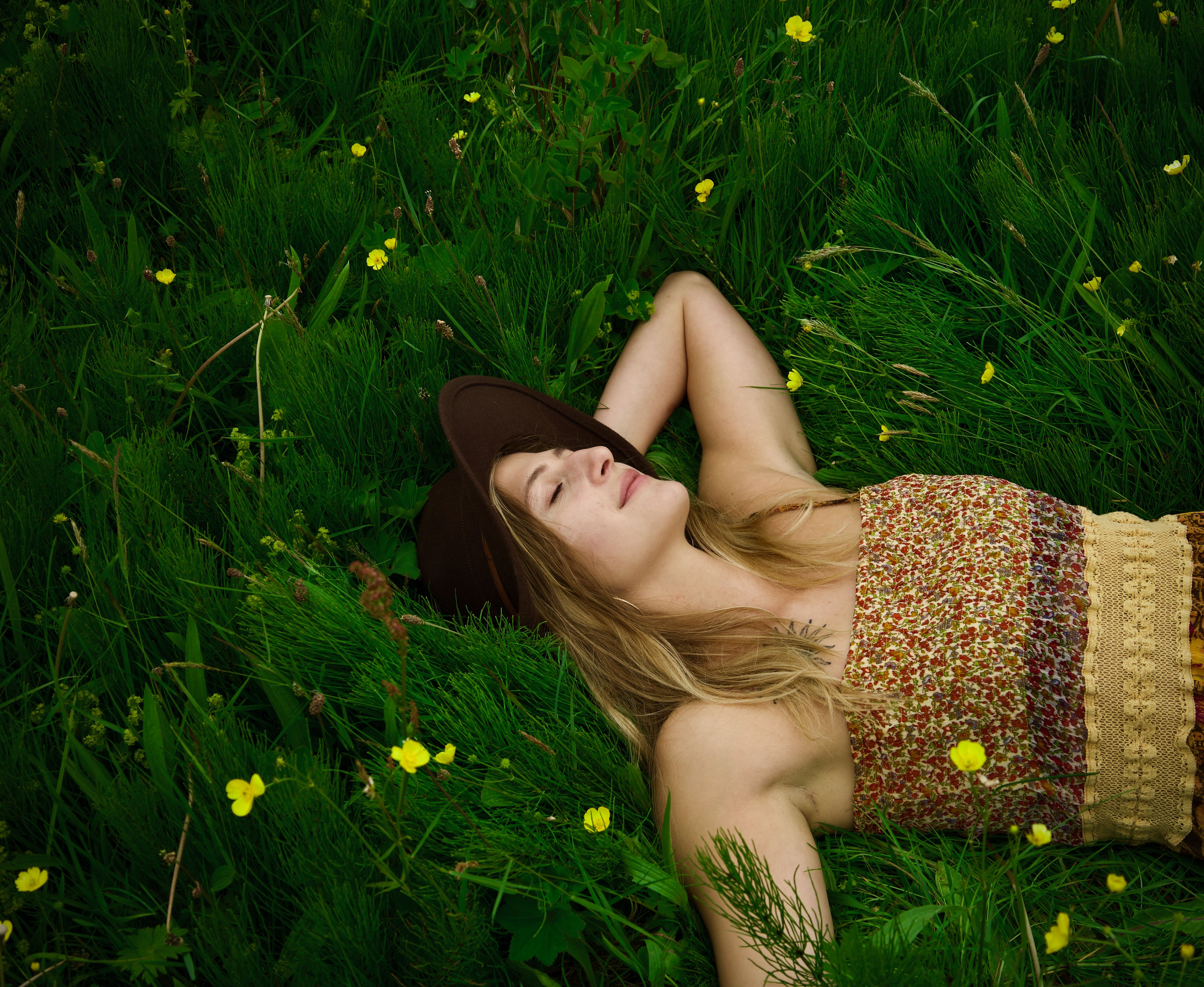Woman lying in green grass with yellow flowers, wearing a floral dress and fedora hat. Peaceful and serene.