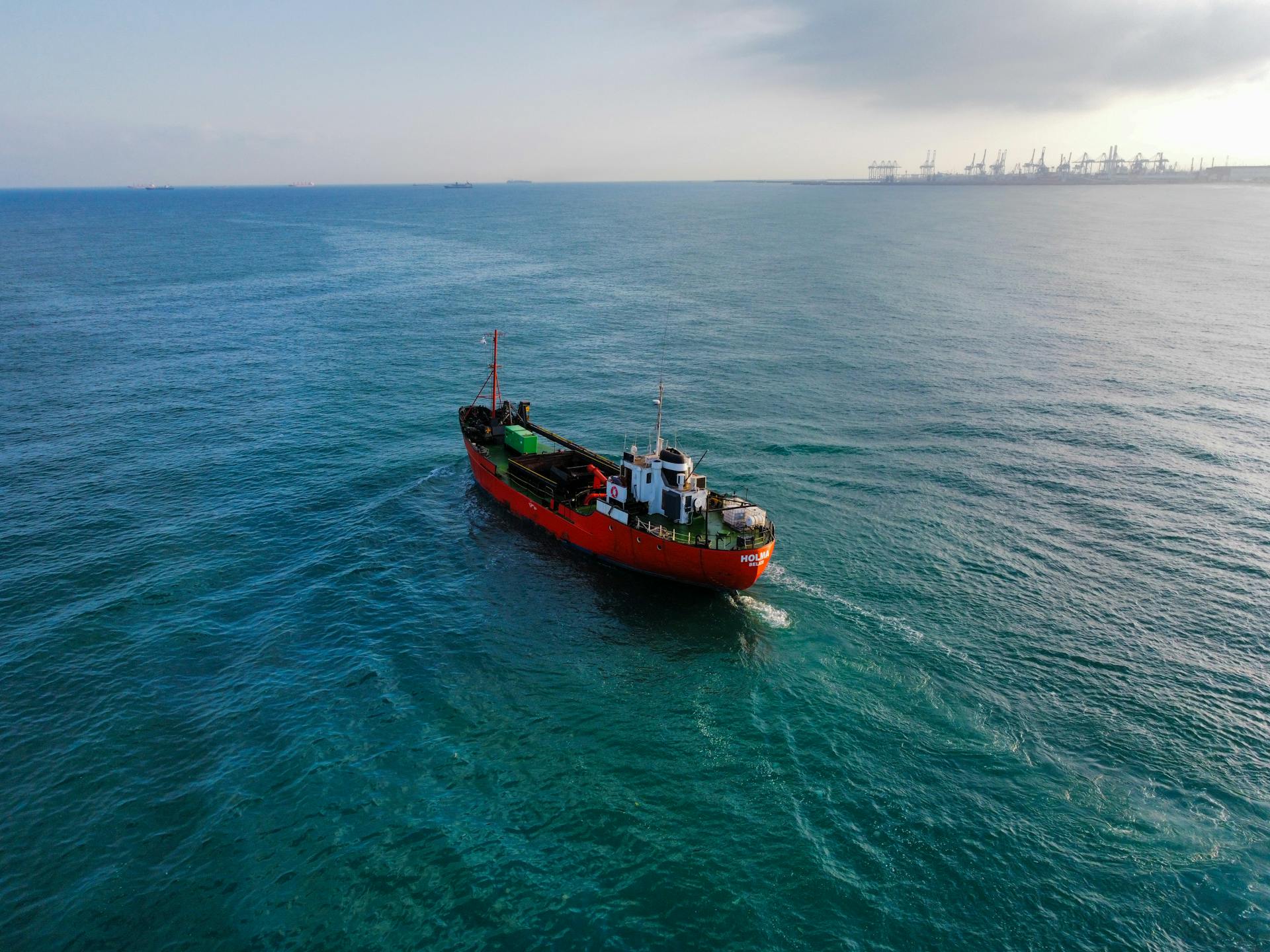 Aerial shot of a cargo ship navigating through tranquil ocean waters near a bustling port.