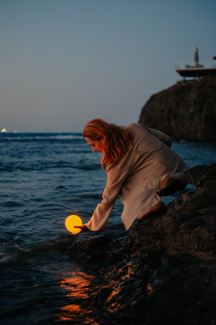 Woman Crouching On Rocks By The Sea At Dusk Holding A Glowing Orb In The Waves