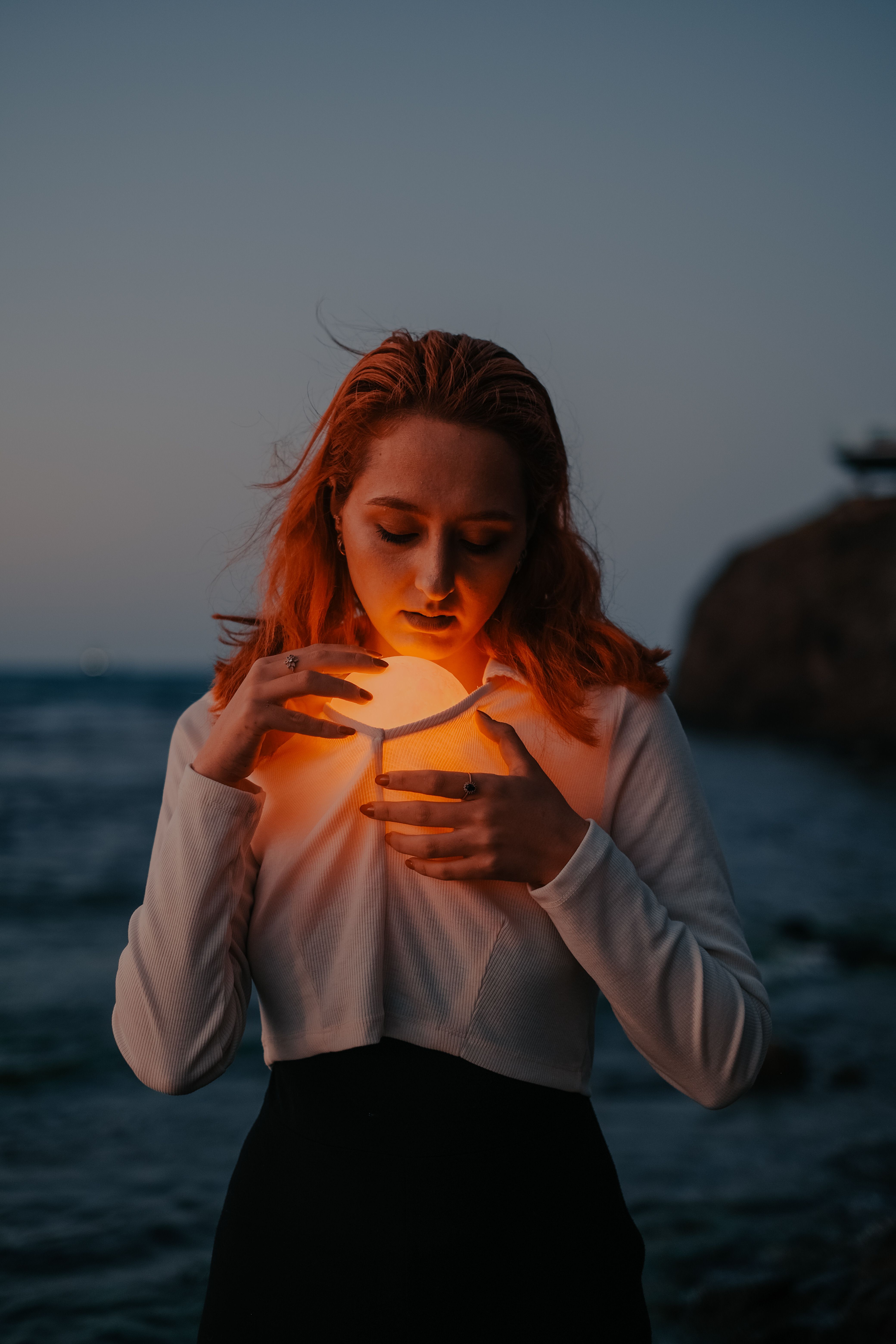 Una mujer serena junto al mar al anochecer, sosteniendo un orbe brillante, creando un ambiente místico.
