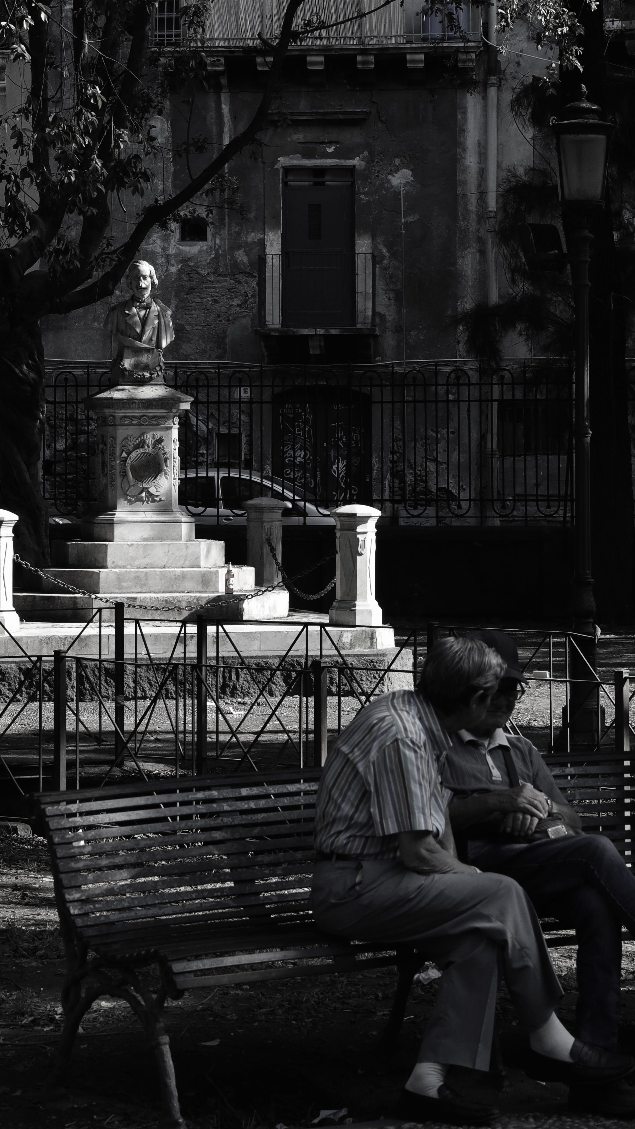 Elderly couple sitting on park bench with statue in background, in monochrome.