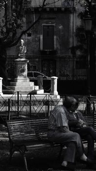 Elderly couple sitting on park bench with statue in background, in monochrome.
