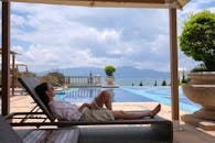A Woman in White Long Sleeves Lying on Brown Wooden Lounge Chair Near Swimming Pool