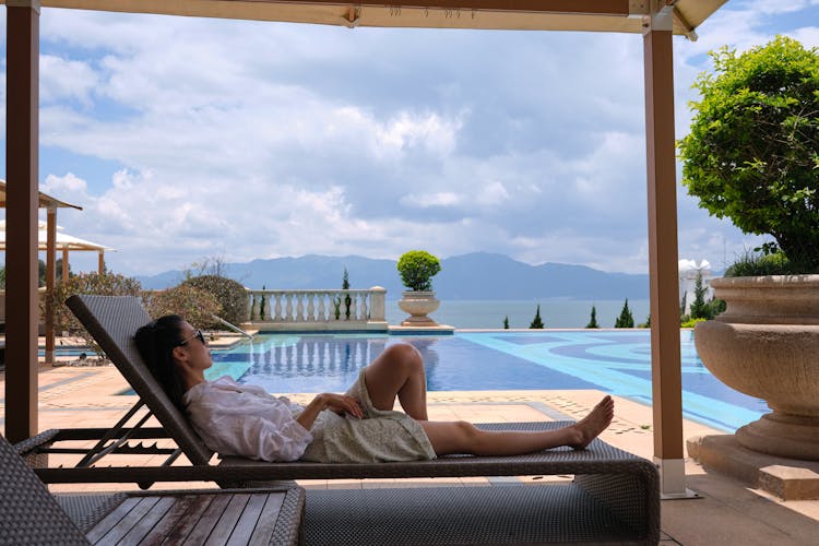 A Woman In White Long Sleeves Lying On Brown Wooden Lounge Chair Near Swimming Pool