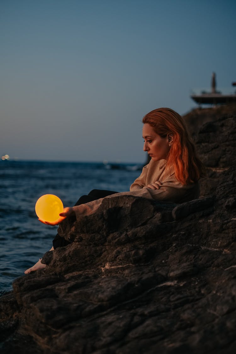 Woman Sitting On A Rock Over Sea At Dusk Holding A Glowing Orb