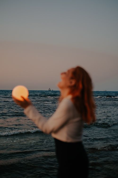 Free A blurred image of a woman holding a glowing orb by the ocean at sunset. Stock Photo