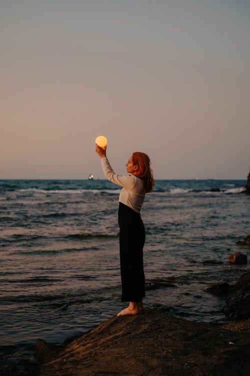 Free Woman standing at the seaside during dusk, holding a glowing orb against the serene sea backdrop. Stock Photo