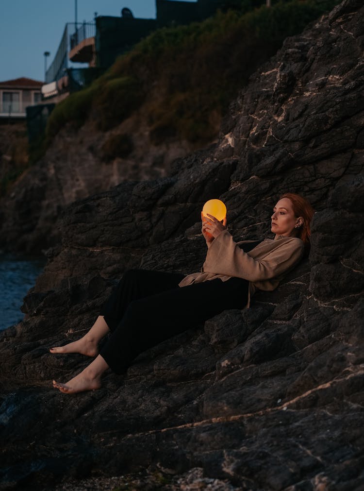 Woman Lying On Rocks By The Sea At Dusk Holding A Glowing Orb