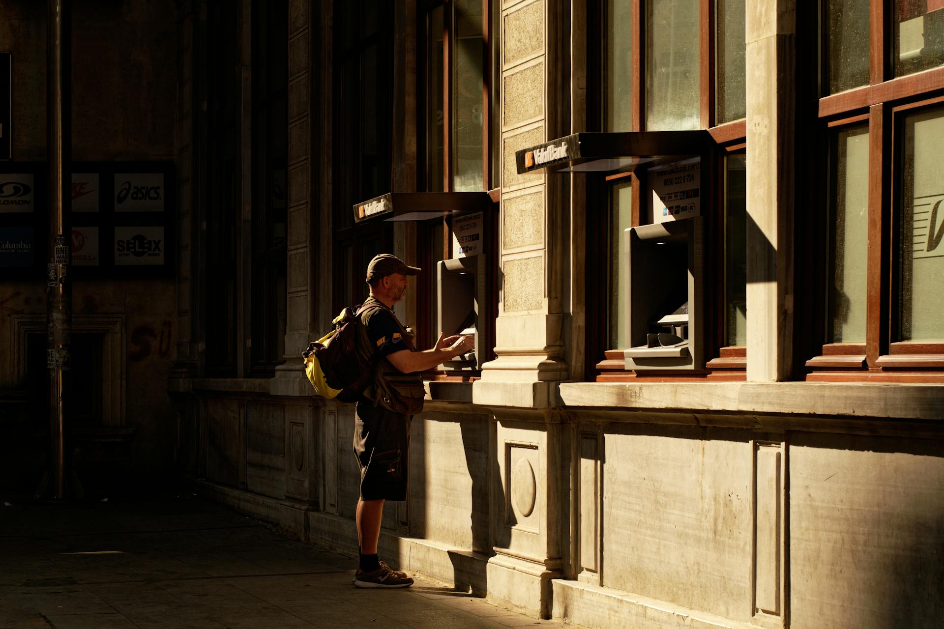 An adult male using an ATM on a historic street corner in warm sunset lighting.