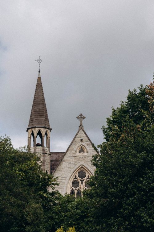 Free Vertical shot of a Gothic church steeple surrounded by trees under a cloudy sky, showcasing religious architecture. Stock Photo