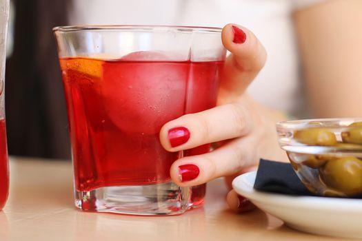 Close-up of a woman's hand holding a red cocktail in a glass with olives on the side.