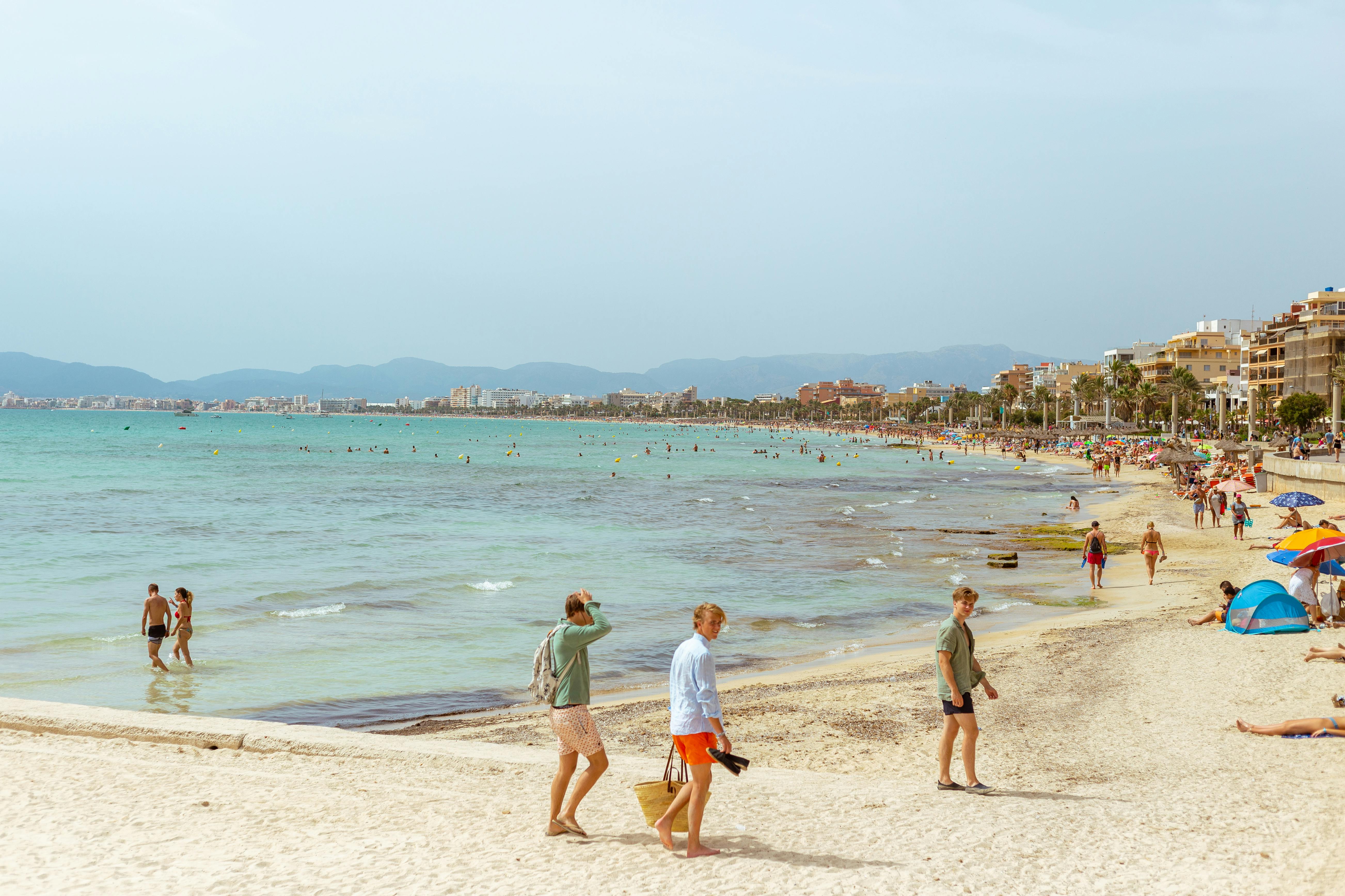 Busy seaside beach with people walking along the shore and swimming, with a coastal town in the distance