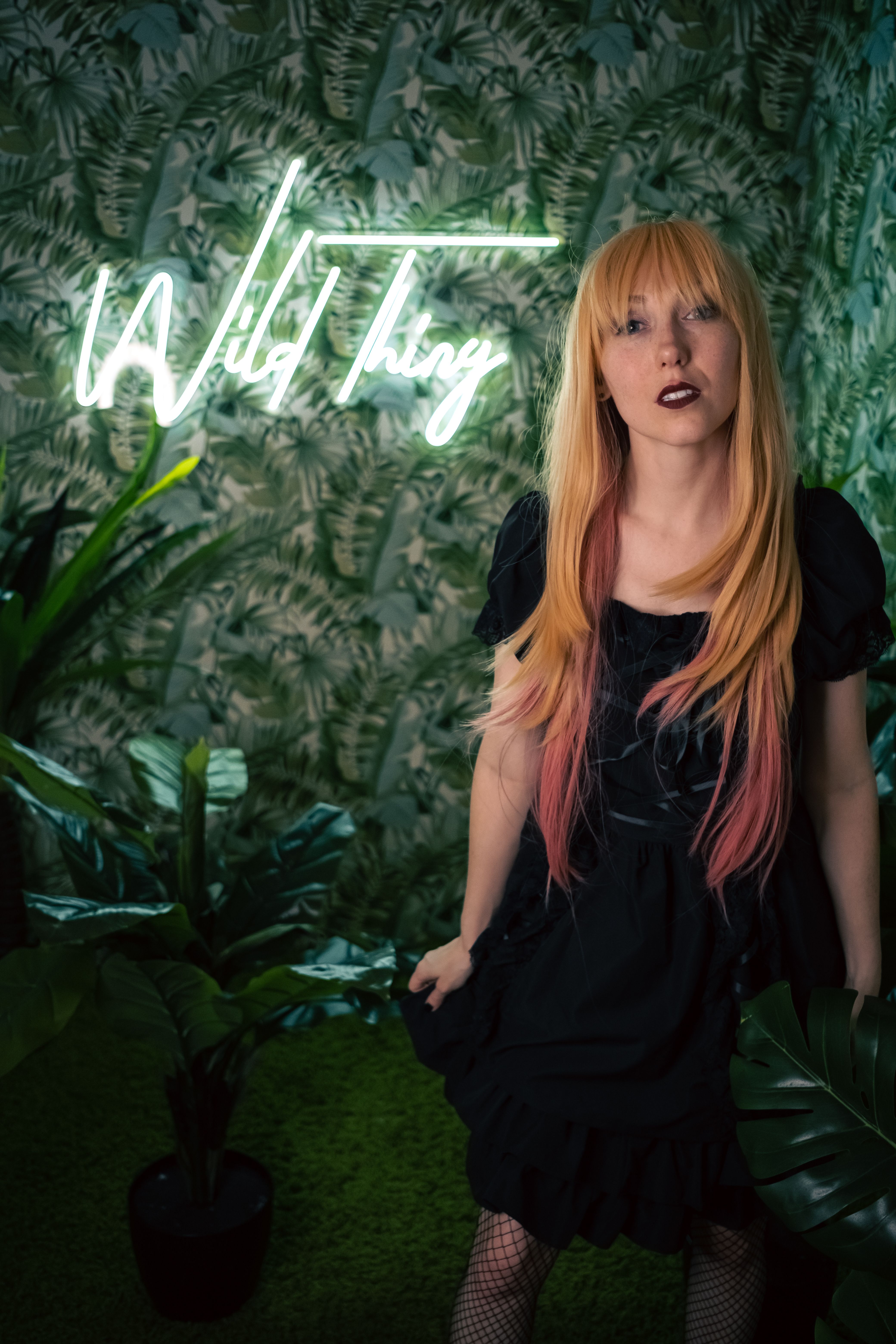 Stylish woman posing in front of neon 'Wild Thing' sign amid lush greenery, exuding vibrant fashion vibes.