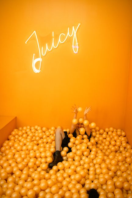 Playful scene with a teenager enjoying an orange ball pit beneath a neon 'Juicy' sign indoors.