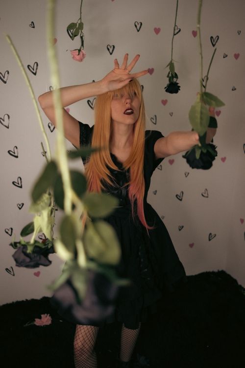 Free A blonde woman in a black dress poses amid hanging roses with artistic heart wall design. Stock Photo