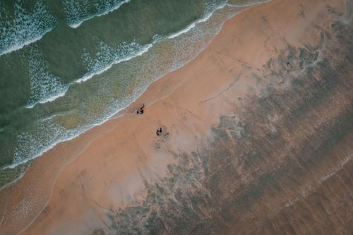 Free Aerial view of a sandy beach with gentle waves and people strolling along the shore. Stock Photo